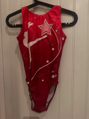 Röd gymnastikdräkt All Star - Snygg röd gymnastikdräkt från All Star Gymnastics med tryck av en gymnast, stjärnor och vita detaljer. Dräkten har ärmlös design och rund halsringning. Materialet är stretchigt och följsamt, perfekt för träning eller tävling inom gymnastik.