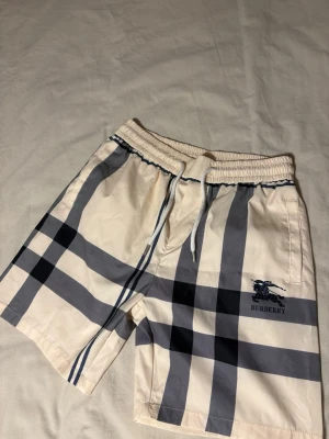 Beige rutiga shorts från Burberry - Snygga beige shorts från Burberry med klassiskt rutigt mönster i svart och grått. Shorts med snörning i midjan och Burberry-logga broderad på ena benet. Perfekta för dig som vill ha en stilren och exklusiv look.