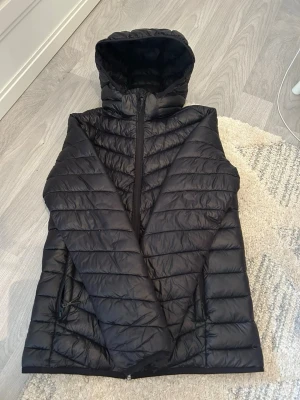 Svart dunväst med huva från Mountain Works - Snygg svart dunväst från Mountain Works med huva och dragkedja framtill. Västen har två sidofickor med dragkedja och ett quiltat mönster. Perfekt för lager-på-lager och håller dig varm under kyliga dagar.