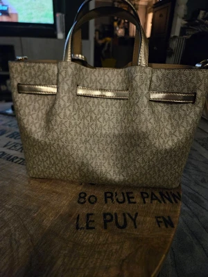 Michael Kors beige handväska - Snygg beige handväska från Michael Kors med guldiga detaljer och klassiskt MK-mönster över hela väskan. Väskan har två handtag, dekorativt MK-hänge och metallicfärgade band. Perfekt för dig som gillar stilrena accessoarer.