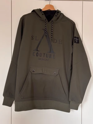 Grön skidjacka från Slade Couture XS - Säljer en grön skidjacka från Slade Couture i storlek XS. Jackan har huva med snörning, stor ficka framtill med tryckknappar och ribbade muddar. Stora logotryck både fram och bak.
