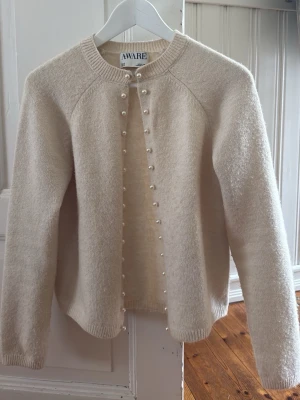 Beige kofta med pärlknappar från AWARE - Supersöt beige kofta från AWARE med rund halsringning och dekorativa pärlknappar längs hela framsidan. Koftan har lång ärm och ribbade muddar vid ärmslut och nederkant. Perfekt för lager på lager och ger en clean, elegant look.