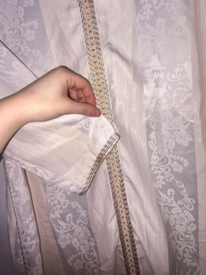 Vit långklänning med gulddetaljer - Elegant vit långklänning med subtilt blommönster och dekorativa guldfärgade band längs framsidan och ärmslut. Klänningen har lång ärm och v-ringad halsringning. Perfekt för dig som vill ha en stilren och klassisk look.