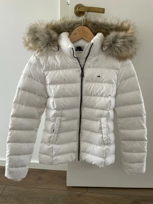 Vit pufferjacka med fuskpäls - Snygg vit pufferjacka från Tommy Hilfiger med avtagbar huva och fuskpäls. Jackan har dragkedja framtill, två sidofickor och diskret logga på bröstet. Perfekt för kalla dagar och ger en clean look.