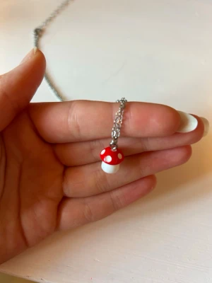 Svampshalsband  - Sött halsband från glitter med en liten röd och vit svampberlock. Kedjan är i silverfärg och har justerbar längd. Perfekt för dig som gillar lekfulla accessoarer och vill addera en unik detalj till din stil.