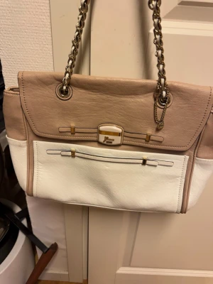 Guess väska  - Snygg handväska från guess  i beige och vitt konstläder med guldfärgade kedjehandtag och detaljer. Väskan har en rektangulär form, dekorativ spänndetalj framtill och en ficka på framsidan. Perfekt accessoar för att lyfta din outfit.
