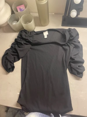 Svart ribbad body från H&M - Snygg svart ribbad topp från H&M med puffärmar och fyrkantig ringning. Toppen är i stretchigt material och har en tight passform som framhäver siluetten. Perfekt att styla med både jeans och kjol för en trendig look.