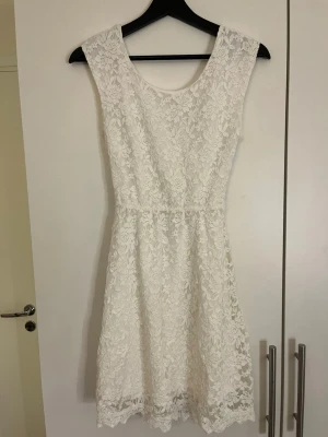 Vit spetsklänning med öppen rygg - Säljer en superfin vit spetsklänning med blommigt mönster. Klänningen är ärmlös, har en kort längd och en snygg öppen rygg med omlottdetalj. .