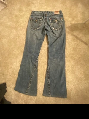 True Religion jeans bootcut - Säljer ett par blå bootcut jeans från True Religion med slitningar och hål på framsidan. Dem är endast använda några gånger så är i bra skick.