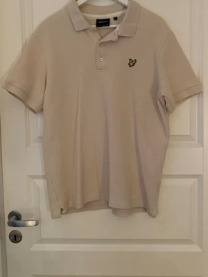 Beige pikétröja från Lyle & Scott - Klassisk beige pikétröja från Lyle & Scott med korta ärmar och krage. Perfekt till jeans eller shorts för en clean och stilren look.