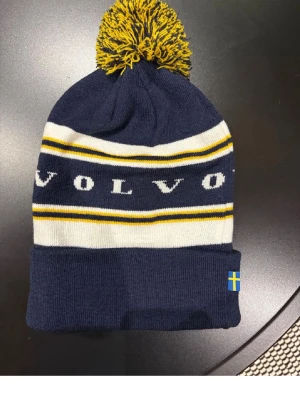 Mörkblå Volvo mössa med tofs - Mörkblå stickad mössa från Volvo med gul och vit rand, stor tofs i gult och blått samt svensk flagga på kanten. Klassisk design med uppvikt kant och tydlig Volvo-logga runt om. Perfekt för kalla dagar och för dig som gillar svensk stil.