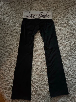 Svarta fold over Love Pink Victoria's Secret - Säljer ett par svarta leggings från Victoria's Secret Pink med grå midja och texten 'Love Pink' i svart. De har bootcut-ben och bred, vikbar midjeresår. Perfekta för en chill och avslappnad stil.