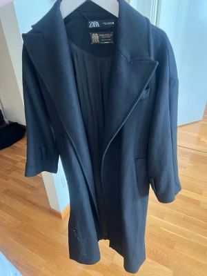 Svart kappa från Zara Manteco - Stilren svart kappa från Zara i samarbete med Manteco, tillverkad av premiumtyg. Kappan har klassisk krage, dolda fickor och rak passform. Perfekt för dig som vill ha en tidlös och elegant look. Fodrad insida för extra komfort.