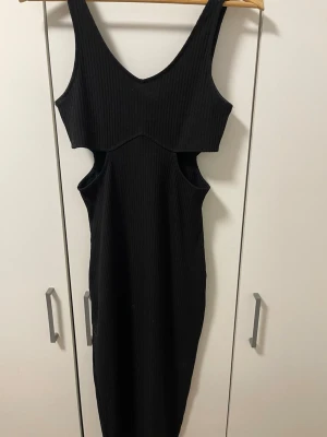 Svart ribbad långklänning med cut-outs - Cool svart långklänning med ribbad struktur och snygga cut-outs på sidorna. Klänningen är ärmlös och har en djup v-ringning. Perfekt för dig som vill sticka ut med en trendig och edgy look.