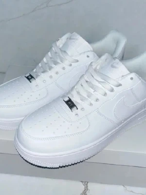 Vita Nike Air Force 1 sneakers - Klassiska helvita Nike Air Force 1 sneakers med låg profil och perforerad tå. Skorna har vit sula, vita skosnören och den ikoniska swoosh-loggan på sidan. Tillverkade i läder för en clean och stilren look.