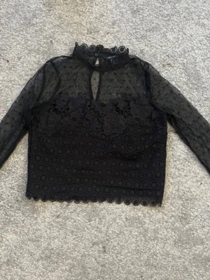 Svart spetsblus med blommor och mesh - Supersnygg svart topp från NA-KD med transparent mesh och prickigt mönster upptill, blommiga spetsdetaljer och långärmad design. Hög hals med knapp i nacken och croppad passform. Perfekt för att lyfta vilken outfit som helst.