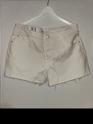 Vita denimshorts från H&M - Vita denimshorts från Bik bok med rå kant nertill och klassisk femficksdesign. Shortsen har knapp och dragkedja framtill samt bälteshällor. Perfekta för varma dagar och enkla att matcha med olika stilar.