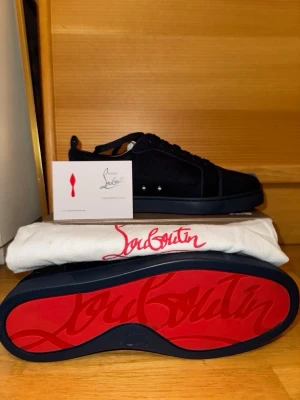 Blåa Louboutin sneakers med röd sula - Säljer ett par blåa sneakers från Christian Louboutin med ikonisk röd sula och diskreta nitar på sidan. Skorna är låga och har en stilren design i mocka med svarta skosnören. Perfekt för dig som vill ha något exklusivt och trendigt.