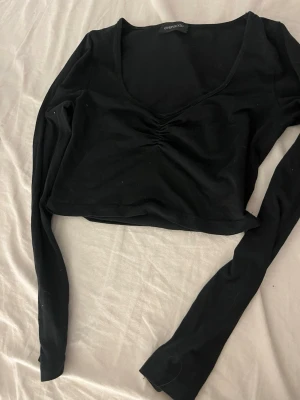 Svart croppad topp med v-ringning - Snygg svart croppad topp från Boohoo med lång ärm och v-ringad hals. Toppen har en lätt rynkning framtill som ger en cool detalj. Tillverkad i mjuk bomullsmix som känns skön mot huden. Perfekt till jeans eller kjol för en trendig look.