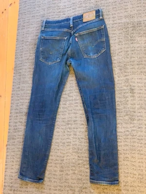 Blå Levi's jeans med raka ben - Säljer ett par klassiska blå Levi's jeans med raka ben och två bakfickor med snygga sömdetaljer. Jeansens material är slitstark denim och de har en läderpatch på linningen. Perfekt för dig som gillar tidlös stil och ikoniska detaljer.