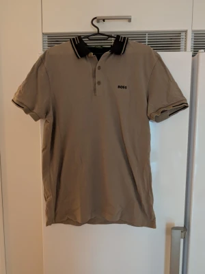 Beige pikétröja från BOSS - Snygg beige pikétröja från BOSS med svart logga broderad på bröstet. Tröjan har klassisk krage med svarta och beige ränder samt knappar framtill. Tillverkad i bomull och har korta ärmar med matchande detaljer på ärmsluten. Slim fit