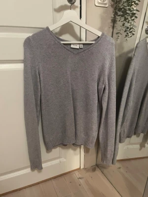Ljusgrå stickad tröja från Vila - Mysig ljusgrå stickad tröja från Vila med v-ringning och långa ärmar. Tröjan har ribbade muddar vid ärmslut och nederkant, vilket ger en snygg och enkel look. Perfekt att matcha med jeans eller kjol för en avslappnad stil.