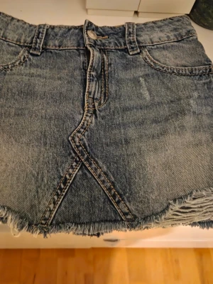 Kort jeanskjol med fransig kant - Snygg kort jeanskjol i blå denim med fransig rå kant nertill och klassiska fickor fram. Kjolen har bälteshällor och dragkedja framtill. Perfekt för dig som gillar en avslappnad och trendig stil.