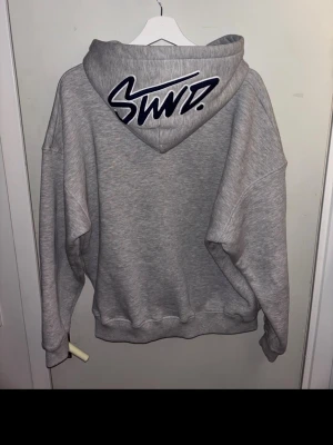 Grå hoodie från Zalando med logga - Säljer en grå hoodie från Zalando med stor svart/vit logga på huvan och mindre logga på bröstet. Hoodien har en klassisk passform, stor känguruficka och ribbade muddar. Materialet är mjukt och bekvämt, perfekt för en avslappnad streetwear-look.