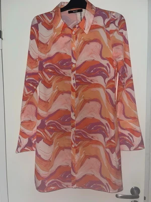 Mönstrad långärmad skjortklänning från Bik Bok - Färgstark skjorta från Bik Bok med abstrakt mönster i orange, rosa, lila och beige toner. Skjortan har klassisk krage, knäppning framtill och långa ärmar. Tillverkad i ett lätt och luftigt material, perfekt för att sticka ut.