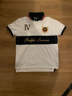 Vit pikétröja från Ralph Lauren - Vit pikétröja från Ralph Lauren med svart krage, broderade detaljer och texten 'Ralph Lauren' i guld över bröstet. Tröjan har korta ärmar, knappar vid halsen och svarta detaljer vid ärmslut och midja. Klassisk och stilren design.