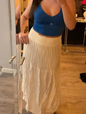 Vit långkjol med resårmidja & linne - Luftig vit långkjol med bred resår i midjan som ger en bekväm passform. Kjolen har ett skirt och lätt material med snyggt skrynklig struktur. Även toppen medföljer