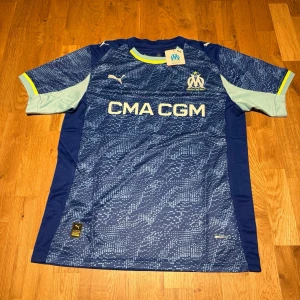 Marseille third kit 25/26  - Säljer en officiell Olympique de Marseille matchtröja från Puma i blått med ljusblå och gula detaljer. Tröjan har klubbmärke och sponsortryck framtill samt korta ärmar. Tillverkad i lätt och ventilerande polyester, perfekt för fotbollsträning.