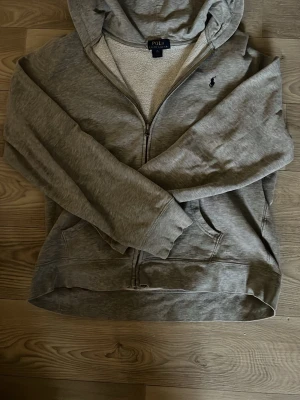 Grå hoodie från Polo Ralph Lauren - Snygg grå zip-hoodie från Polo Ralph Lauren med huva och fickor framtill. Klassisk liten mörkblå logga på bröstet och mjukt, fleecefodrat innerfoder. Perfekt för en avslappnad och stilren look. Storlek 18-20 vilket motsvarar S ungefär. Lite lös i midjan men inget som påverkat mig. Annars är det en klockren tröja. Hör av er vid frågor eller om ni vill ha mer bilder!