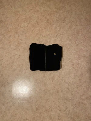 Svart zip hoodie från Lyle & Scott - Säljer en svart hoodie med dragkedja från Lyle & Scott. Klassisk design med huva, två fickor framtill och den ikoniska gula örnloggan broderad på bröstet. Perfekt för en avslappnad och stilren look. Mjuk bomullsblandning för extra komfort.
