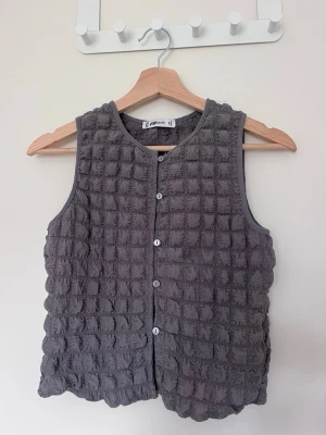 Grå topp med knappar - Grå väst med quiltad struktur och rutigt mönster. Toppen är ärmlös, har rund halsringning och stängs med knappar framtill.