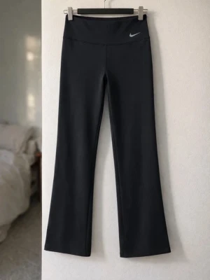 Svarta träningsbyxor från Nike - Svarta träningsbyxor från Nike med utsvängda ben och hög midja. Byxorna har en diskret Nike-logga framtill och är tillverkade i ett stretchigt material som passar perfekt för träning eller gym. Snygg och enkel design som är lätt att matcha.