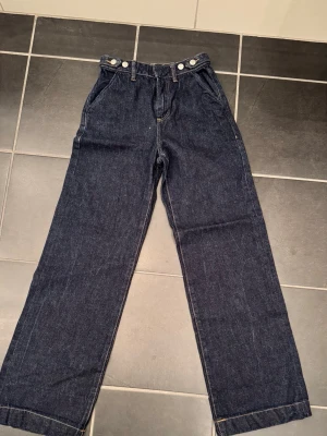 Mörkblå raka jeans med hög midja - Säljer ett par mörkblå jeans med raka ben och hög midja. Jeansen har klassisk femficksdesign, bälteshällor och silverfärgade knappar i midjan. Materialet är kraftig denim. Perfekta för en avslappnad och trendig look.