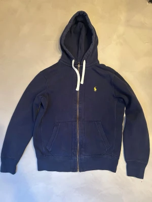 Mörkblå zip up från Polo Ralph Lauren - Mörkblå zip up från Polo Ralph Lauren med dragkedja, vit dragsko och klassisk gul logga på bröstet. Tillverkad i mjukt bomullsmaterial och har kängurufickor framtill. Perfekt för en avslappnad och stilren look. Nypris är ca 2600kr, priset går att diskutera. Hör av er vid frågor.