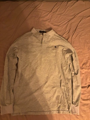 Grå långärmad pikétröja Polo Ralph Lauren - Klassisk grå långärmad pikétröja från Polo Ralph Lauren med liten broderad logga på bröstet. Tröjan har krage, knäppning vid halsen och ribbade muddar vid ärmsluten. Perfekt för en clean och stilren look.