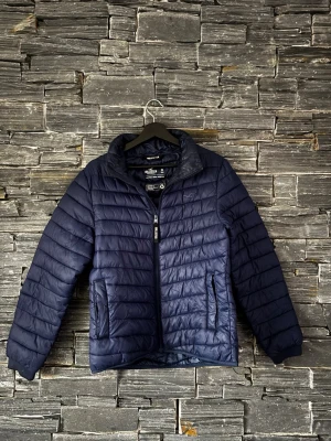 Mörkblå pufferjacka från Walkman - Snygg mörkblå pufferjacka från Hollister med quiltad design och dragkedja framtill. Jackan har två fickor med dragkedja och hög krage som skyddar mot kyla. Perfekt för kalla vinterdagar och enkel att matcha med olika outfits.