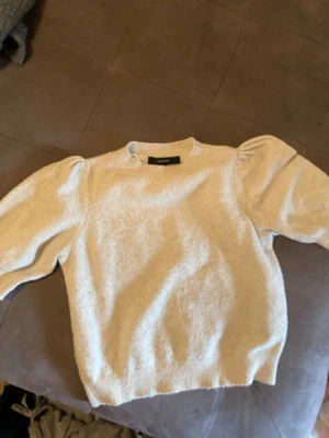 Beige stickad tröja Vero Moda - Snygg beige stickad tröja från Vero Moda med puffärmar och ribbade muddar vid hals, ärmslut och nederkant. Tröjan har en mjuk struktur och rund halsringning, perfekt för en trendig och avslappnad look.