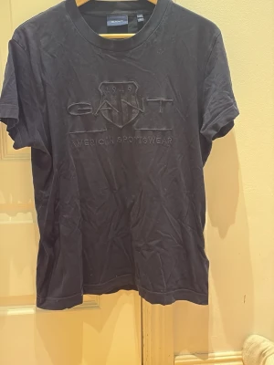 Svart GANT t-shirt med broderad logga - Snygg svart t-shirt från GANT med broderad logga och texten 'American Sportswear' framtill. Klassisk rund hals och korta ärmar. Tillverkad i mjuk bomull som känns skön mot huden. Perfekt till jeans eller shorts för en clean och enkel stil.