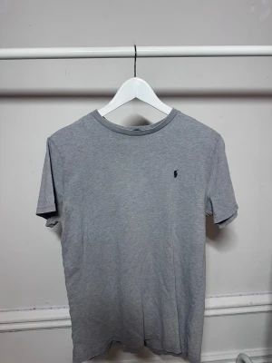 Grå t-shirt från Polo Ralph Lauren - Snygg grå t-shirt från Polo Ralph Lauren med klassisk rund hals och den ikoniska mörkblå broderade loggan på bröstet. Tillverkad i mjukt bomullstyg som känns skönt mot huden. Perfekt basplagg för en clean och stilren look. Hör av dig vid frågor eller funderingar eller vill ha fler bilder🙌