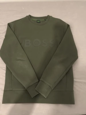 Hugo Boss sweatshirt  - Snygg olivgrön sweatshirt från BOSS med diskret logga i samma färg på bröstet. Tröjan har rund halsringning, långa ärmar och ribbade muddar. Perfekt för en stilren och avslappnad look. Materialet känns mjukt och bekvämt mot huden. Tröjan är knappt använd och allt funkar med den som den ska, den är i storlek S. nypris 1500
