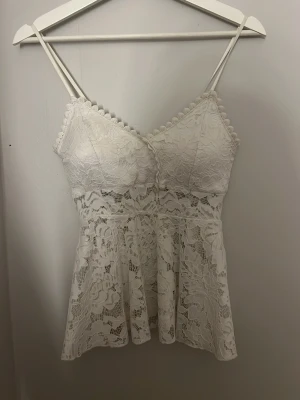 Vit spetslinnetopp med smala band - Supersöt vit linnetopp med tunna axelband och vacker spets över hela toppen. Toppen har en lätt utsvängd passform och dekorativ kant vid bysten. Perfekt för dig som gillar romantisk och feminin stil.