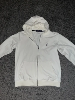 Polo Ralph Lauren hoodie - Säljer en clean vit hoodie från Polo Ralph Lauren med dragkedja och huva. Klassisk logga broderad på bröstet i mörkblått. Hoodien har två fickor framtill och ribbade muddar. Perfekt för en chill och stilren look.