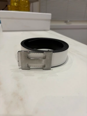 Vändbart svart och vitt skärp med H-spänne - Snyggt vändbart skärp i svart och vitt med ett stort, stilrent H-format metallspänne. Perfekt accessoar för att lyfta din outfit och matcha olika looks. Skärpet har rena linjer och är gjort i slätt skinn.