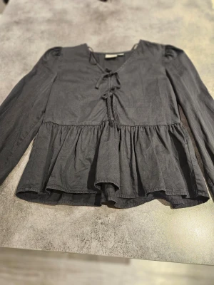 Gina tricot  - Snygg svart blus med långa ärmar, v-ringning och dekorativ snörning framtill. Blusen har volang nedtill. Har haft den cirka 3-5 gånger. Typ som ny. Storlek 146-152 