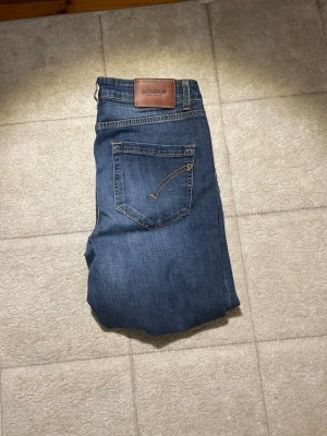 Blå jeans från Dondup - Säljer ett par klassiska blå jeans från Dondup med raka ben och snygga detaljer på bakfickan. Jeansen har fem fickor, gylf med knapp och dragkedja samt en läderpatch med logga bak. Materialet är denim i bomull och färgen är mörkblå.