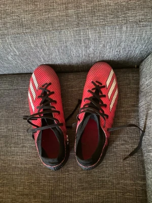 Röda Adidas fotbollsskor med dobbar - Säljer ett par röda Adidas fotbollsskor med klassiska vita ränder på sidan och svarta skosnören. Skorna har en syntetisk ovandel med struktur för bättre bollkontroll och rosa dobbsula för spel på gräs. Perfekta för dig som vill sticka ut på planen!